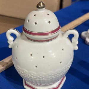May include: White ceramic jar with a lid, featuring a gold finial and a red stripe. The jar has small holes and decorative detailing. It has two handles and a hexagonal base with a red trim. A vintage decorative piece.