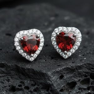 Può includere: Un paio di orecchini a lobo a forma di cuore. Ogni orecchino presenta un centro di pietra preziosa rosso intenso, circondato da un alone di piccole pietre chiare. Gli orecchini sono incastonati in un metallo color argento. Lo sfondo è una superficie scura e strutturata.