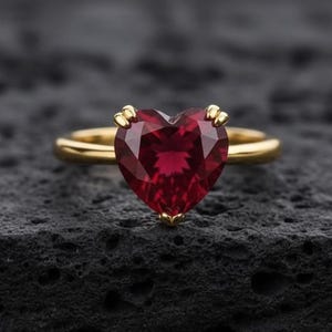 Peut inclure: Une bague en or avec une pierre précieuse rouge foncé en forme de cœur et facettée. La bague est posée sur une surface sombre et texturée, mettant en valeur la couleur et le design. L'anneau de la bague est en or poli simple.
