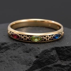 Könnte beinhalten: Ein goldener Ring mit drei ovalen Edelsteinen in Rot, Grün und Blau. Der Ring hat ein verziertes Design mit Schnörkeln zwischen den Edelsteinen. Der Ring liegt auf einer dunklen, strukturierten Oberfläche.