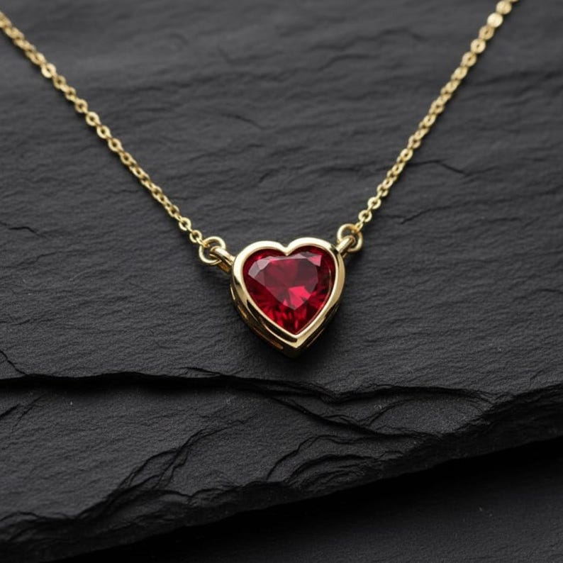 May include: A gold-colored necklace with a heart-shaped, red gemstone pendant. The pendant is set in a gold bezel and hangs from a delicate gold chain. The necklace is displayed on a dark, textured surface.