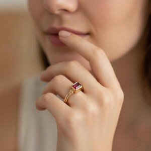 May include: A gold ring featuring a rectangular, deep pink gemstone set in a bezel setting. The band of the ring is smooth and polished, showcasing a simple design that highlights the vibrant color and clarity of the central stone.