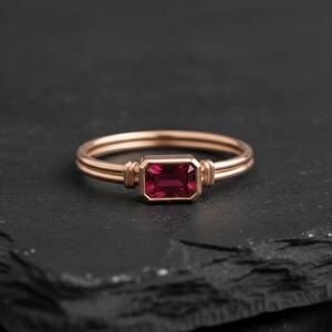 May include: A rose gold ring featuring a rectangular, emerald-cut deep red gemstone set in a bezel setting. The band consists of two parallel thin wires that meet at the stone, which is flanked by small vertical metallic accents on each side.