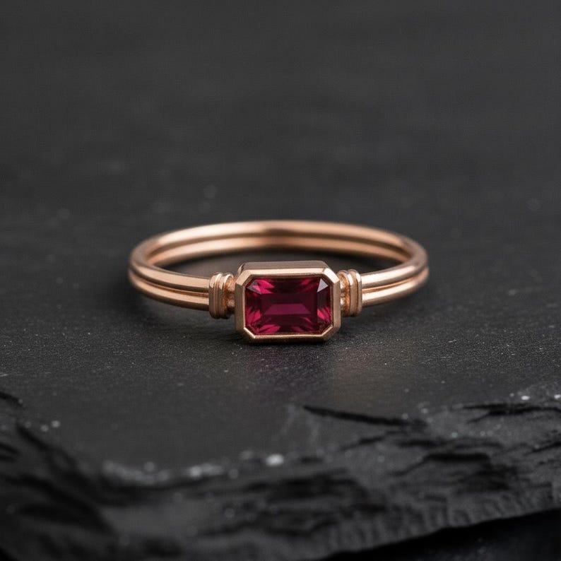 May include: A rose gold ring featuring a rectangular, emerald-cut deep red gemstone set in a bezel setting. The band consists of two parallel thin wires that meet at the stone, which is flanked by small vertical metallic accents on each side.