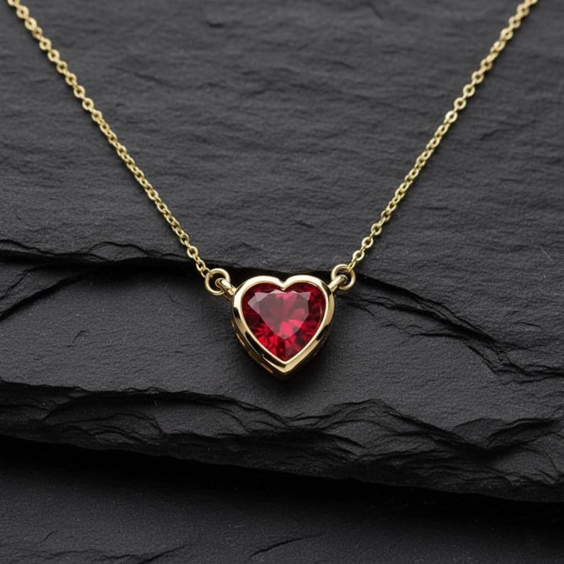 May include: A gold-colored necklace with a heart-shaped, red gemstone pendant. The pendant is set in a gold bezel and hangs from a delicate chain. The necklace is displayed on a black, textured surface.