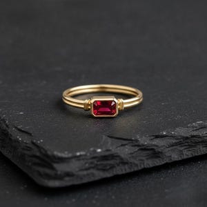 May include: A gold ring featuring a rectangular, emerald-cut red gemstone set in a bezel setting. The band consists of two thin, parallel gold wires that meet at the stone, which is flanked by small, vertical decorative gold bars on each side.