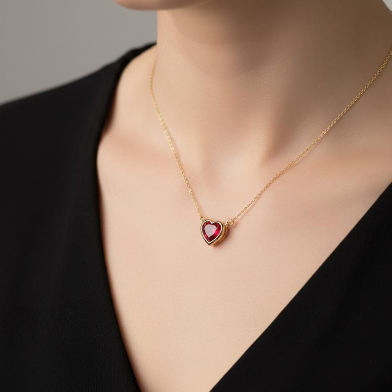 May include: A gold necklace featuring a heart-shaped, ruby-red gemstone pendant. The pendant is set in a gold bezel and hangs from a delicate gold chain. The necklace is worn against a black top.