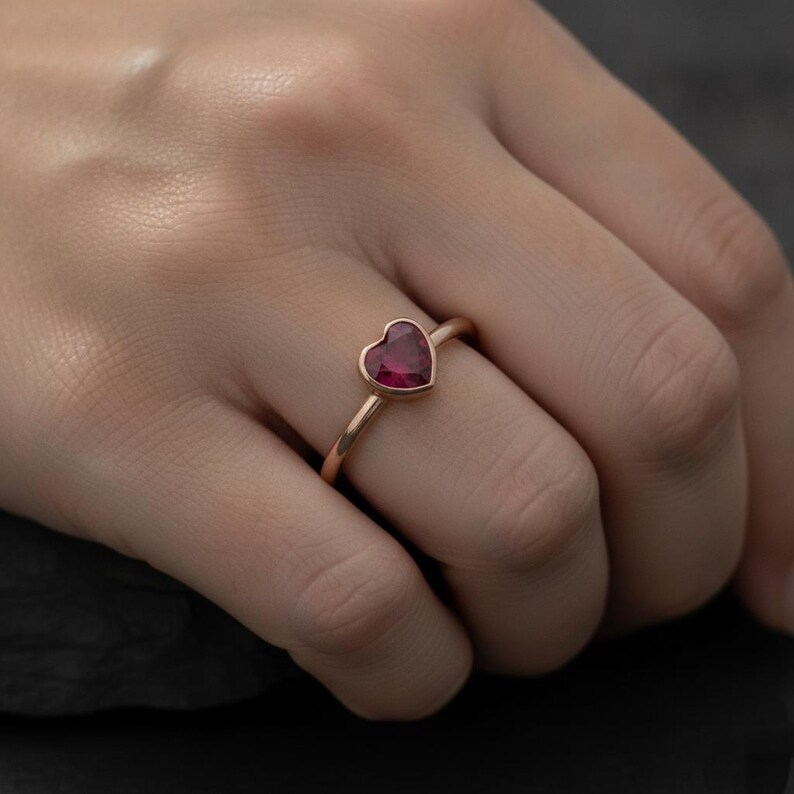 Puede incluir: Un anillo con una piedra preciosa de color rosa intenso en forma de coraz&oacute;n, montada en una banda de oro rosa pulido. La piedra est&aacute; asegurada en un engaste de bisel, creando un perfil suave y continuo en el dedo. La banda es fina y redondeada.
