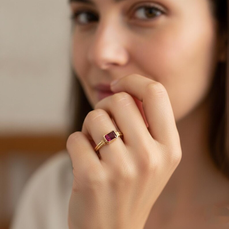 May include: A gold ring featuring a rectangular, deep pink gemstone set in a bezel setting. The band has a polished finish and a simple, clean design that highlights the vibrant color and geometric shape of the central stone.