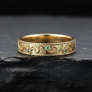 Puede incluir: Un anillo dorado con un patrón floral grabado y tres pequeñas piedras preciosas verdes. El diseño detallado del anillo se destaca sobre un fondo oscuro, resaltando el brillo de las piedras y la artesanía.
