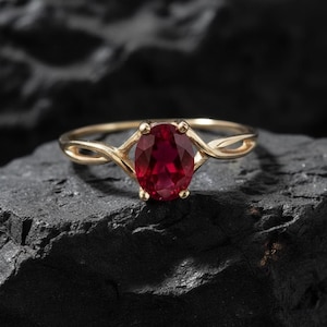 May include: A gold ring featuring a large, oval-cut ruby gemstone. The ruby is a deep red hue, set in a four-prong setting. The ring's band has a twisted design. The ring is presented on a dark, textured surface.