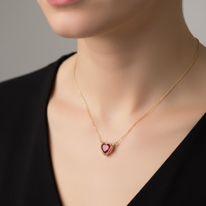 May include: A gold-colored necklace featuring a heart-shaped pendant with a vibrant red gemstone. The pendant is set in a gold frame and hangs from a delicate gold chain. The necklace is displayed on a person wearing a black top.