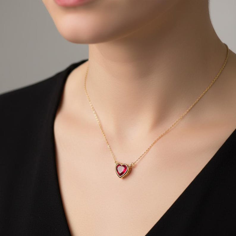 May include: A gold-colored necklace featuring a heart-shaped pendant with a vibrant red gemstone. The pendant is set in a gold frame and hangs from a delicate gold chain. The necklace is displayed on a person wearing a black top.