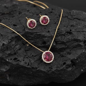 Op de afbeelding: Een goudkleurige sieradenset met een hangerketting en bijpassende oorstekers. Elk stuk heeft een grote, ronde, dieprode edelsteen omringd door een halo van kleinere, heldere stenen. De set wordt gepresenteerd op een donkere, getextureerde ondergrond.