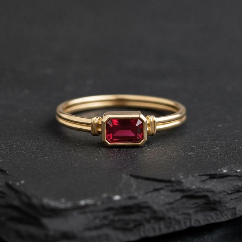 May include: A gold ring featuring a rectangular, emerald-cut red gemstone set in a bezel setting. The band consists of two thin, parallel gold wires that meet at the stone, which is flanked by small, decorative gold ridges on either side.