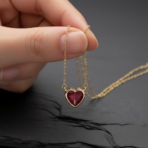 May include: A gold-colored necklace with a heart-shaped, red gemstone pendant. The pendant is set in a gold bezel and hangs from a delicate gold chain. The necklace is displayed against a dark background.