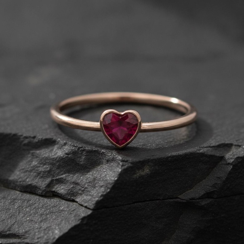 Puede incluir: Un anillo fino de oro rosa con una peque&ntilde;a piedra preciosa de color rojo intenso en forma de coraz&oacute;n engastada en el centro. El anillo tiene un acabado met&aacute;lico liso y pulido, y la piedra est&aacute; facetada, reflejando la luz en su forma.