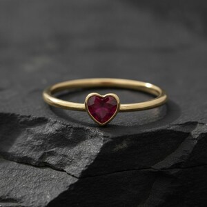 Puede incluir: Un anillo de banda fina de oro con una peque&ntilde;a piedra preciosa de color rojo intenso en forma de coraz&oacute;n engastada en el centro. El anillo tiene un acabado liso y pulido, y la piedra est&aacute; facetada, reflejando la luz en su superficie.