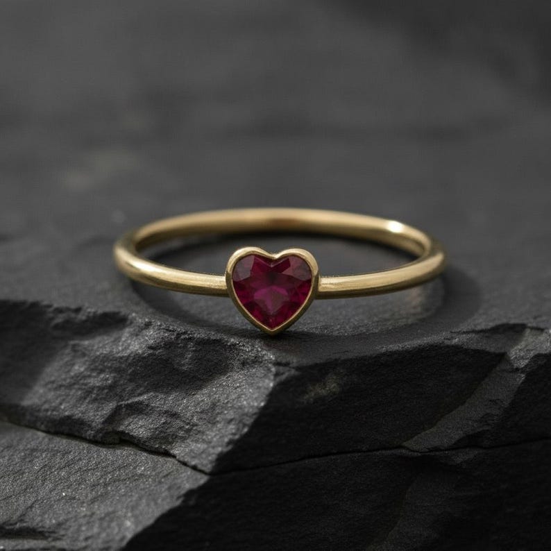 Puede incluir: Un anillo de banda fina de oro con una peque&ntilde;a piedra preciosa de color rojo intenso en forma de coraz&oacute;n engastada en el centro. El anillo tiene un acabado liso y pulido, y la piedra est&aacute; facetada, reflejando la luz en su superficie.