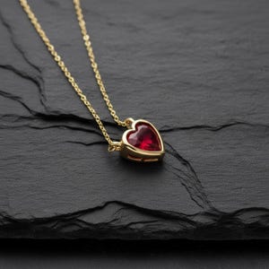 May include: A gold-colored necklace with a heart-shaped, red gemstone pendant. The pendant is set in a gold bezel and hangs from a delicate gold chain. The necklace is displayed on a dark, textured surface.