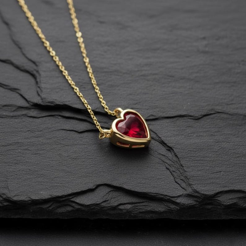 May include: A gold-colored necklace with a heart-shaped, red gemstone pendant. The pendant is set in a gold bezel and hangs from a delicate gold chain. The necklace is displayed on a dark, textured surface.