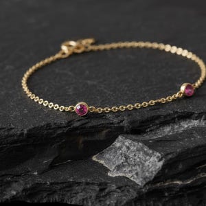May include: A delicate gold bracelet with a chain design. The bracelet features two round, vibrant pink gemstones. The bracelet is displayed on a dark, textured surface, highlighting the jewelry's elegant design.