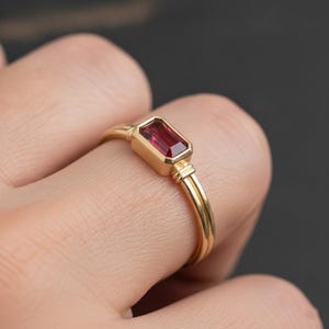 May include: A gold ring featuring a rectangular, emerald-cut deep red gemstone set in a bezel setting. The band is thin and smooth, connecting to the stone setting with a small, double-bar detail on each side of the central red gemstone.