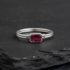 May include: A silver-toned ring featuring a rectangular, emerald-cut deep pink gemstone set in a bezel setting. The band consists of two parallel thin silver wires that connect to the central stone setting with small, ribbed metal accents.