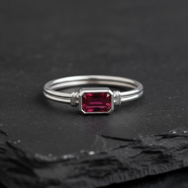 May include: A silver-toned ring featuring a rectangular, emerald-cut deep pink gemstone set in a bezel setting. The band consists of two parallel thin silver wires that connect to the central stone setting with small, ribbed metal accents.