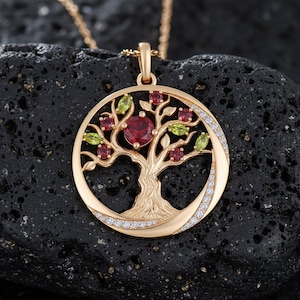 Op de afbeelding: Een goudkleurige hangerketting met een levensboomontwerp. De boom heeft een centrale rode edelsteen, rode en groene accenten en een rand van kleine witte edelstenen. De ketting wordt gepresenteerd op een zwart, getextureerd oppervlak.