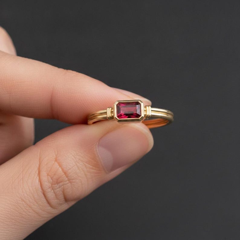 May include: A gold ring featuring a rectangular, emerald-cut deep red gemstone set in a bezel setting. The band consists of two parallel gold wires that meet at the stone, creating a structured, layered appearance with a polished metallic finish.