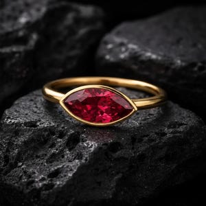 May include: A gold ring with a marquise-cut, deep red gemstone. The ring is set in a gold bezel and rests on a dark, textured surface. The gemstone's shape is elongated, and the gold band is smooth and polished.