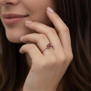 Puede incluir: Un anillo fino de oro con una piedra preciosa de color rosa oscuro en forma de coraz&oacute;n, montada en un engaste de bisel. El aro es liso y met&aacute;lico, con un dise&ntilde;o sencillo que resalta la piedra central en forma de coraz&oacute;n en el dedo.