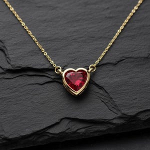May include: A gold necklace with a heart-shaped, ruby-red gemstone pendant. The pendant is set in a gold bezel and hangs from a delicate gold chain. The necklace is displayed on a black surface.