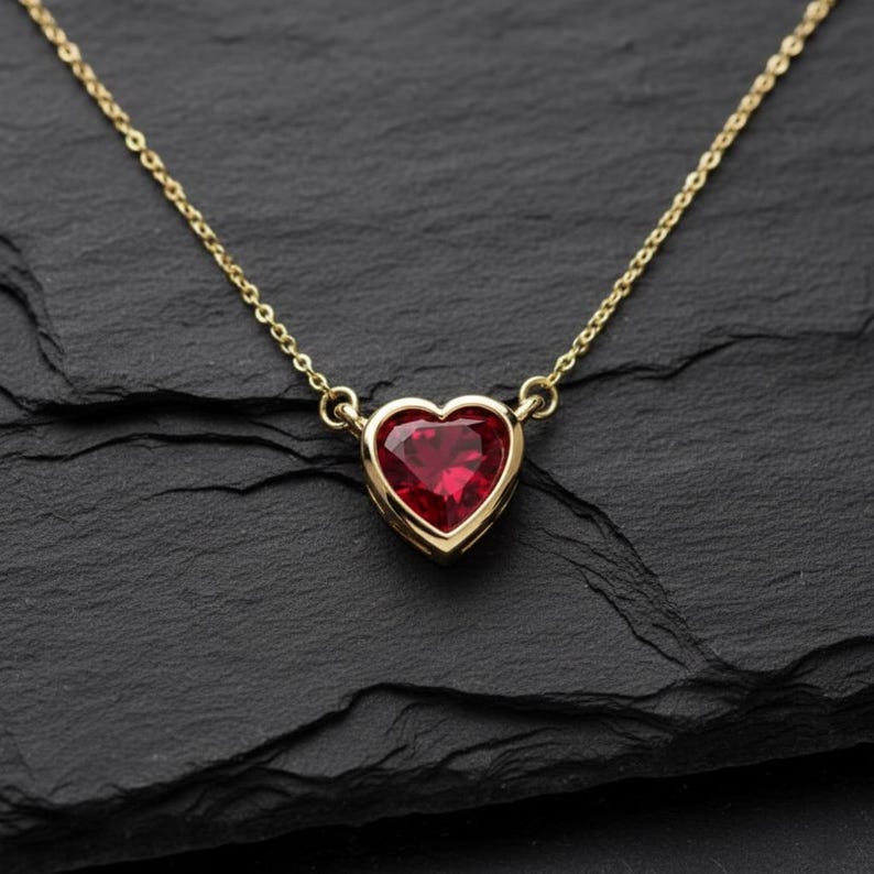 May include: A gold necklace with a heart-shaped, ruby-red gemstone pendant. The pendant is set in a gold bezel and hangs from a delicate gold chain. The necklace is displayed on a black surface.