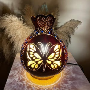 Peut inclure: Lampe en forme de gourde rouge foncé avec un motif de papillon. La lampe présente un papillon sculpté au centre, avec de petits points bleus et un dessus festonné. La lampe projette une ombre en forme de papillon.