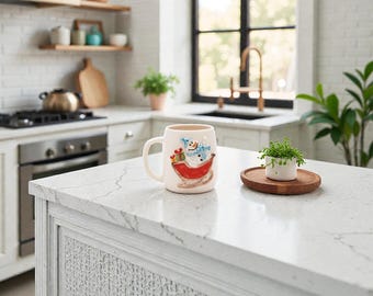 May include: A cream-colored ceramic mug with a snowman design in a red sleigh, holding gifts. The mug has a large handle and is on a white countertop in a kitchen setting. A small plant sits on a wooden tray.