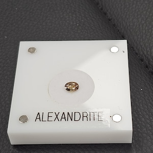 May include: An oval-shaped alexandrite gemstone, displayed on a white square holder with the word "ALEXANDRITE" printed on it. The gemstone is a light brown color and is set in a clear, circular setting. The holder has four silver-colored metal studs.