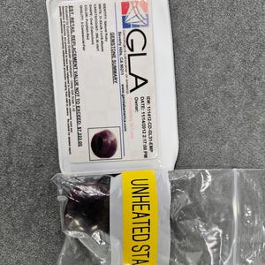 Puede incluir: Un rubí natural, de forma oval cabujón, color rojo púrpura, acompañado de una tarjeta de identificación del Gemological Laboratory of America (GLA). La tarjeta detalla las medidas del rubí, 3,18x2,91x1,85cm, y el peso, 206,35 quilates. Una etiqueta amarilla dice "UNHEATED STAR RUBY."