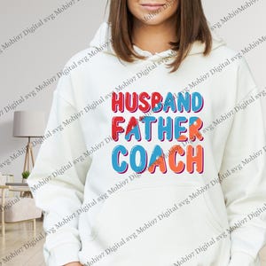May include: Cream-coloured hoodie featuring the words "HUSBAND FATHER COACH" in red, blue, and orange block letters. The text is stacked vertically. A comfortable, casual item of clothing.