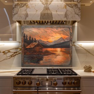 Op de afbeelding: Een ingelijst landschapsschilderij met een hut aan een meer bij zonsondergang. Het kunstwerk gebruikt een warm kleurenpalet van oranje, rood en bruin, met bergen en bomen op de achtergrond. Het schilderij is gemonteerd boven een roestvrijstalen gasfornuis.