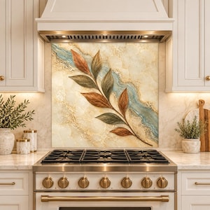 May include: A square kitchen backsplash panel featuring a central branch with brown and dark green leaves. The background displays a cream-colored marble texture with a diagonal vein of light blue and gold accents, positioned above a stainless steel stove.