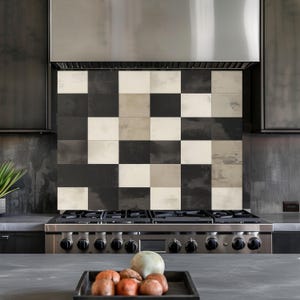 May include: A kitchen backsplash with a chequered pattern of black, white, and grey square tiles. The tiles are above a stainless steel gas hob. A stainless steel cooker hood is above the tiles.