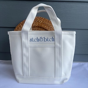 Może przedstawiać: Biała pł&oacute;cienna torba typu tote z niebieskim haftem "stch&btch". Torba ma prostokątny kształt z małą kieszonką z przodu i dwoma uchwytami. Wewnątrz torby znajduje się tkany, okrągły przedmiot.