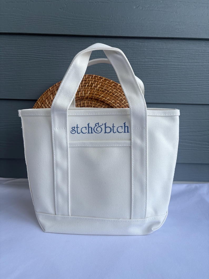 Może przedstawiać: Biała pł&oacute;cienna torba typu tote z niebieskim haftem "stch&btch". Torba ma prostokątny kształt z małą kieszonką z przodu i dwoma uchwytami. Wewnątrz torby znajduje się tkany, okrągły przedmiot.