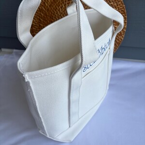 Może przedstawiać: Biała pł&oacute;cienna torba typu tote z solidnymi uchwytami i małą kieszonką. Torba ma niebieski haftowany tekst na kieszeni. Torba znajduje się przed tkanym wiklinowym tłem.