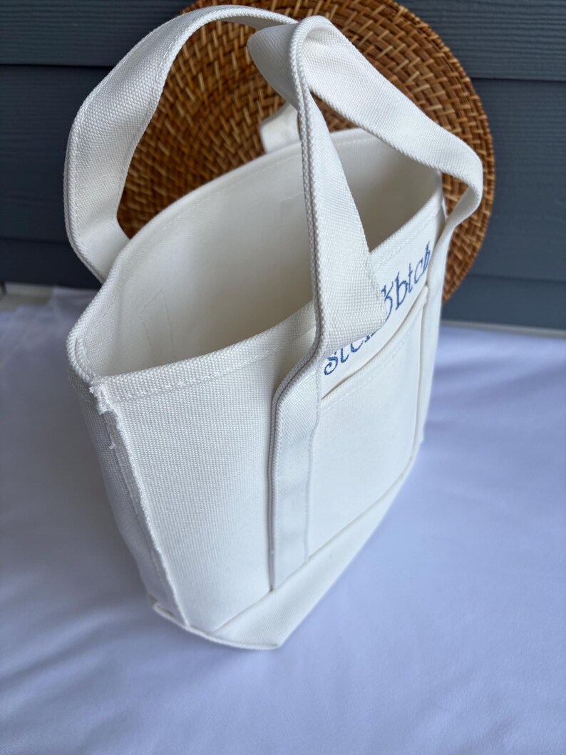 Może przedstawiać: Biała pł&oacute;cienna torba typu tote z solidnymi uchwytami i małą kieszonką. Torba ma niebieski haftowany tekst na kieszeni. Torba znajduje się przed tkanym wiklinowym tłem.