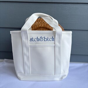 Może przedstawiać: Biała pł&oacute;cienna torba z dwoma uchwytami i niebieskim haftem "stch&btch". Torba jest prostokątna i wydaje się pusta. Wewnątrz torby widoczny jest tkany przedmiot.