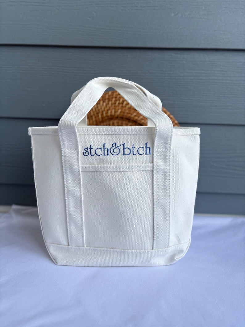 Może przedstawiać: Biała pł&oacute;cienna torba z dwoma uchwytami i niebieskim haftem "stch&btch". Torba jest prostokątna i wydaje się pusta. Wewnątrz torby widoczny jest tkany przedmiot.