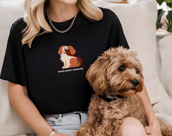 Könnte beinhalten: Eine Frau trägt ein schwarzes T-Shirt mit einem Comic-Hund und dem Text "Cookie Quality Inspector". Sie hält einen braunen und weißen Hund auf ihrem Schoß.